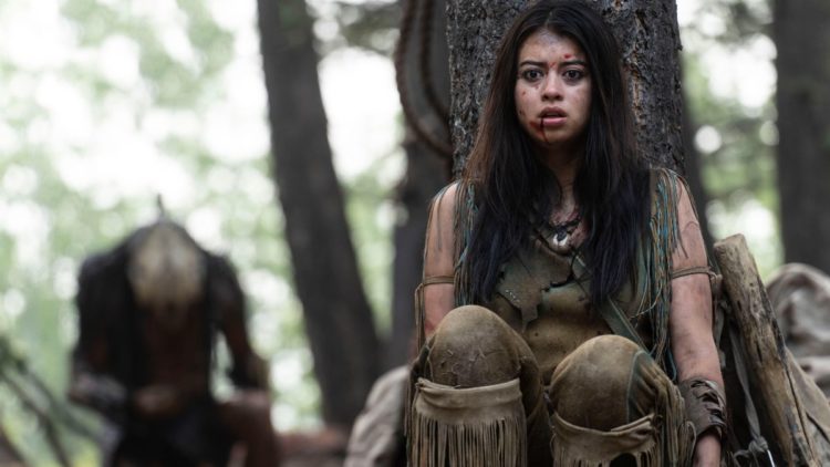 Amber Midthunder as Naru in Prey fighting Predator in the forest with the Predator behind in Prey.