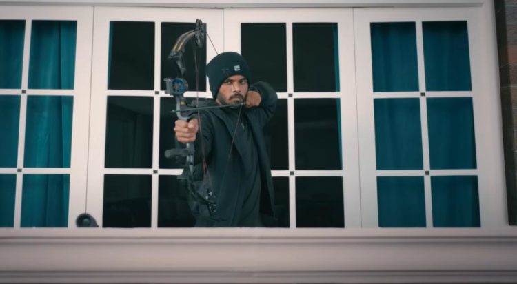 A man dressed all in black draws a modern archery bow standing on a balcony