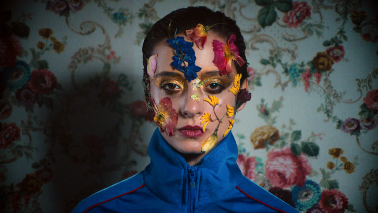 Angela Bundalovic as Miu stands with flowers on her face in a blue track suit