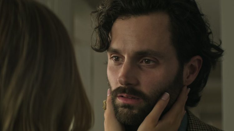 Penn Badgley as Joe Goldberg standing at a window in a brown sweater in You