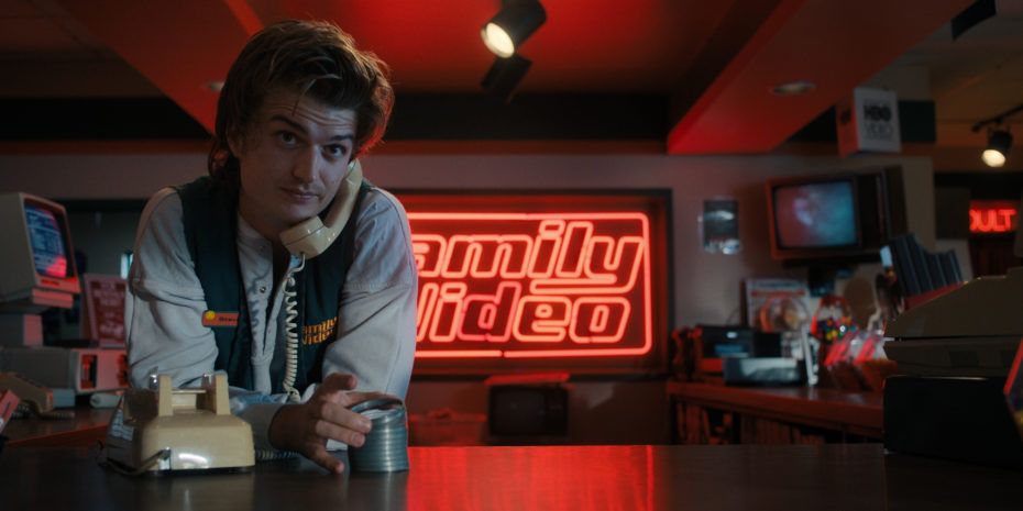 Joe Keery as Steve Harrington working at Family Video with neon sign in background while he's on phone in Stranger Things