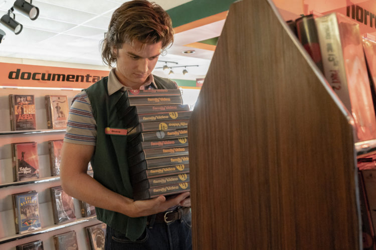 Joe Keery as Steve Harrington holding stack of videos wearing vest in Stranger Things